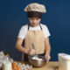 Isolated portrait of cute teenage boy learning how to make cookies at culinary workshop, holding whisker while beating eggs, standing at kitchen table with all ingredients and utensils on it